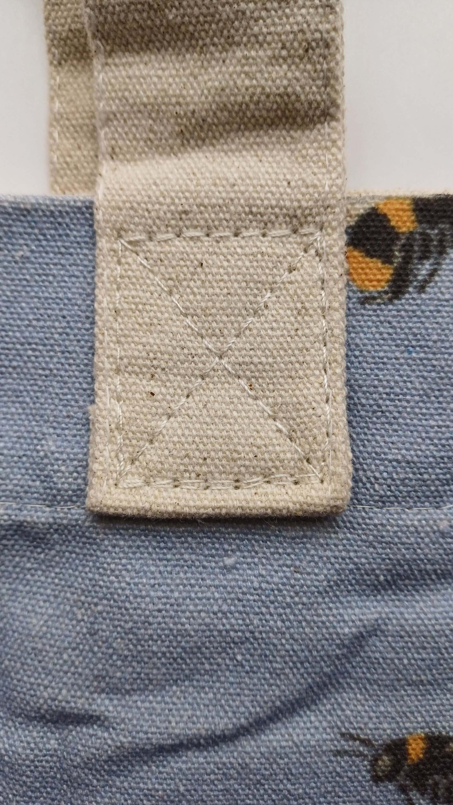 Close-up of a sturdy cream canvas handle, reinforced with stitching, attached to a light blue bee-printed tote bag. Durable construction.