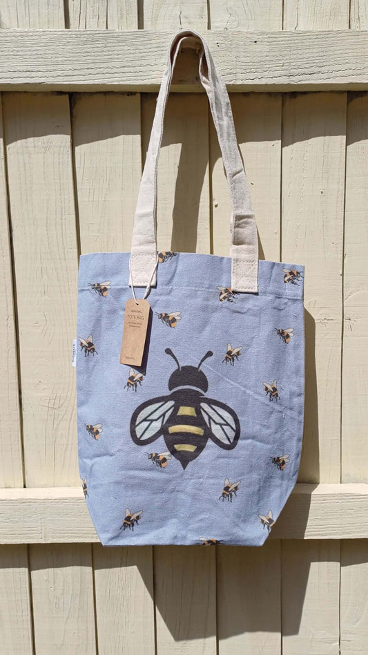 Light blue canvas tote featuring a large central bee and smaller scattered bees. Includes an interior pocket and sturdy cream-colored straps.
