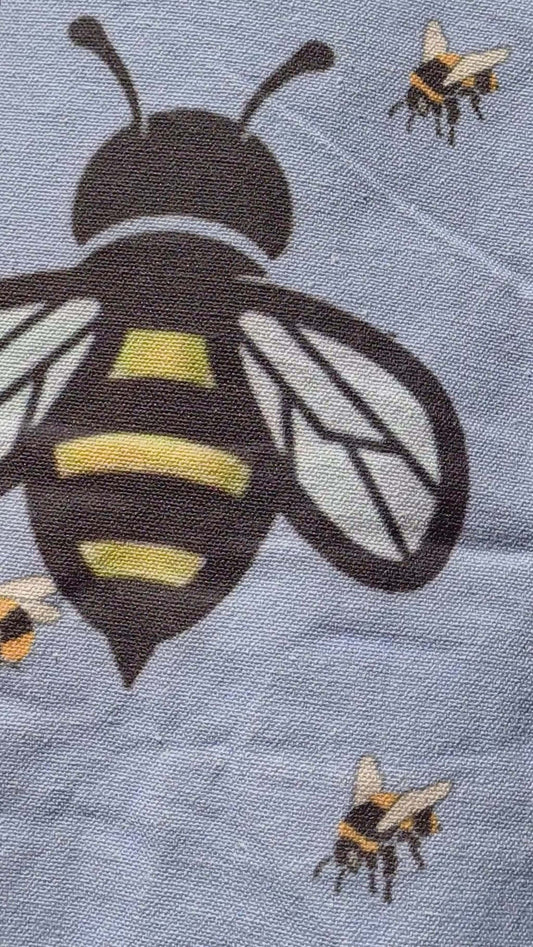 Close-up of a light blue canvas tote bag displaying detailed bee illustrations in black and yellow. Textured fabric.