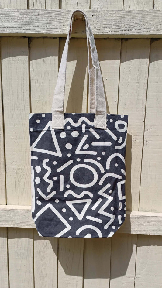 Black canvas tote bag featuring a bold white graffiti doodle print. Includes a convenient inner pocket and sturdy cream-colored handles.