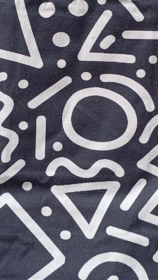 Close-up of a black canvas tote bag's pattern: bold white graffiti-style shapes including circles, triangles, lines, and squiggles.