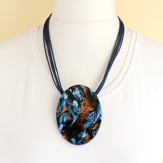 Bold blue pendant necklace with a striking abstract pattern in black and orange. Adjustable cord for varied lengths.
