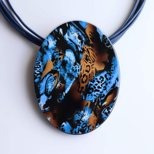 Striking oval pendant necklace with a bold abstract design in blue, black, and orange animal print. Hung on multiple blue cords.