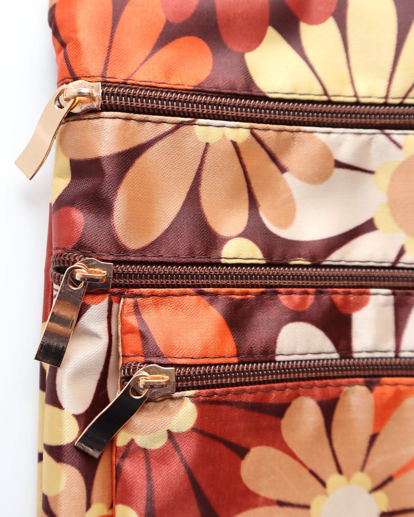 Close-up of a retro floral crossbody bag showing three zippered compartments with gold-toned pulls. Warm brown fabric with orange and cream daisies.