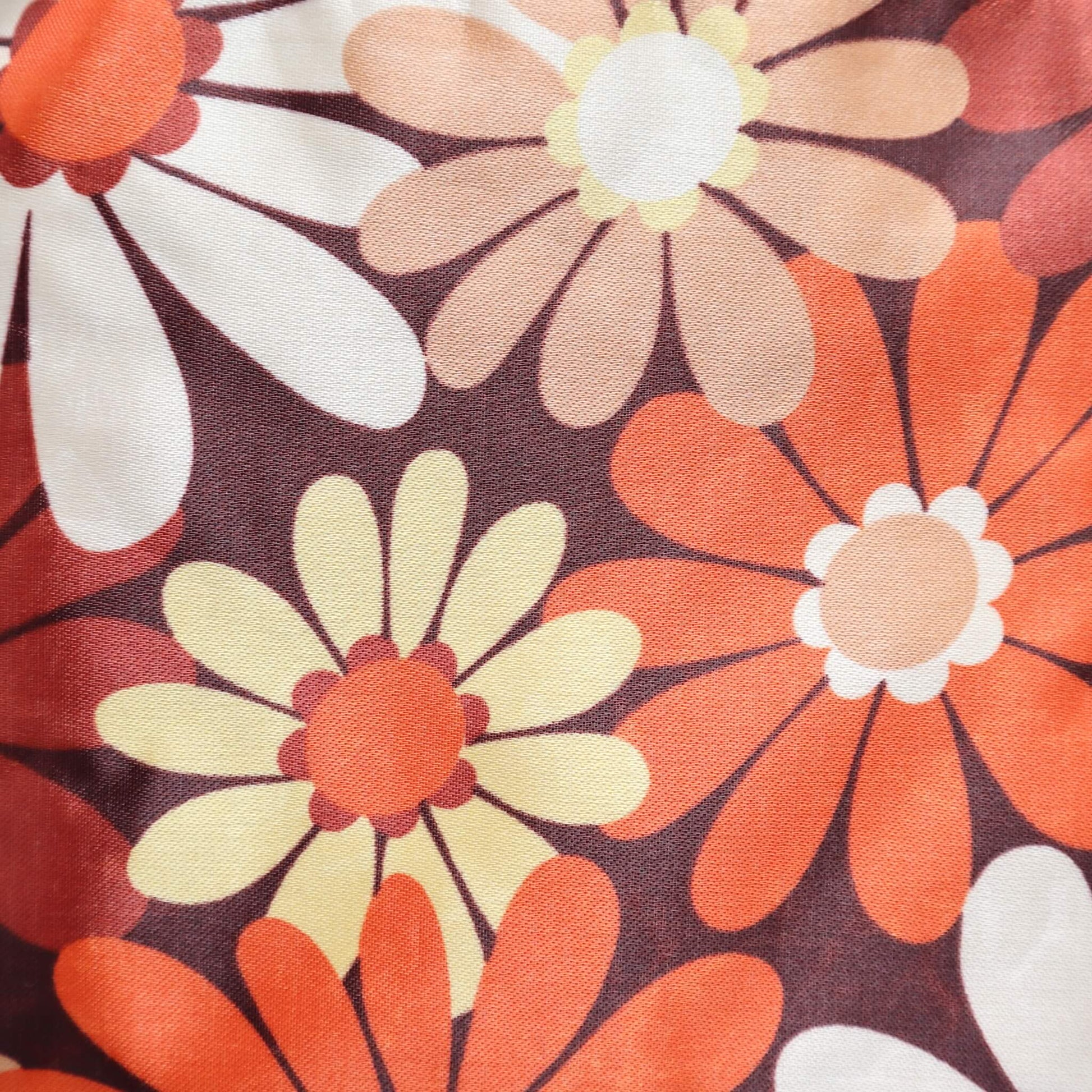 Close-up of a crossbody bag's fabric: a retro daisy chain print in shades of brown, orange, cream, and yellow. Detailed floral pattern.