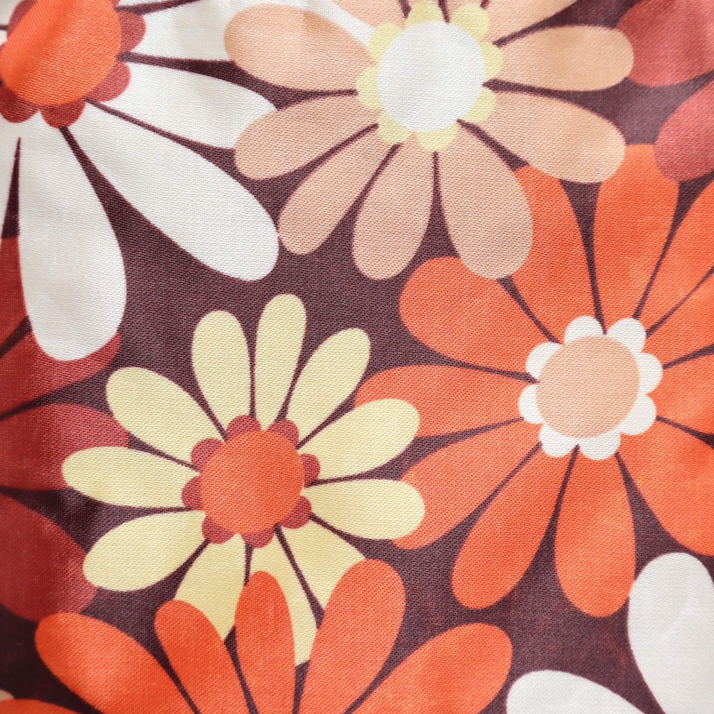 Close-up of a crossbody bag's fabric: a retro daisy chain print in shades of brown, orange, cream, and yellow. Detailed floral pattern.