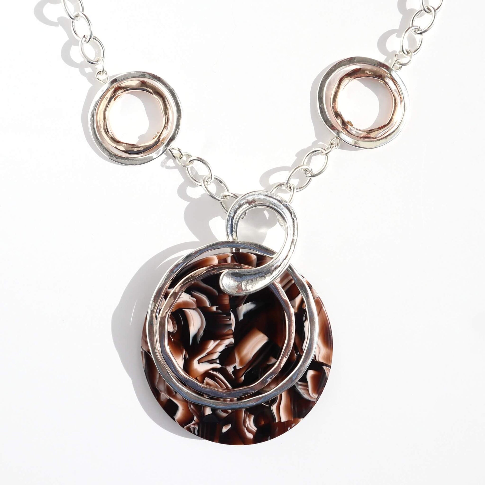 Silver-toned chain necklace with silver and rose gold tone open rings, and a large round pendant with a swirling brown and cream pattern.