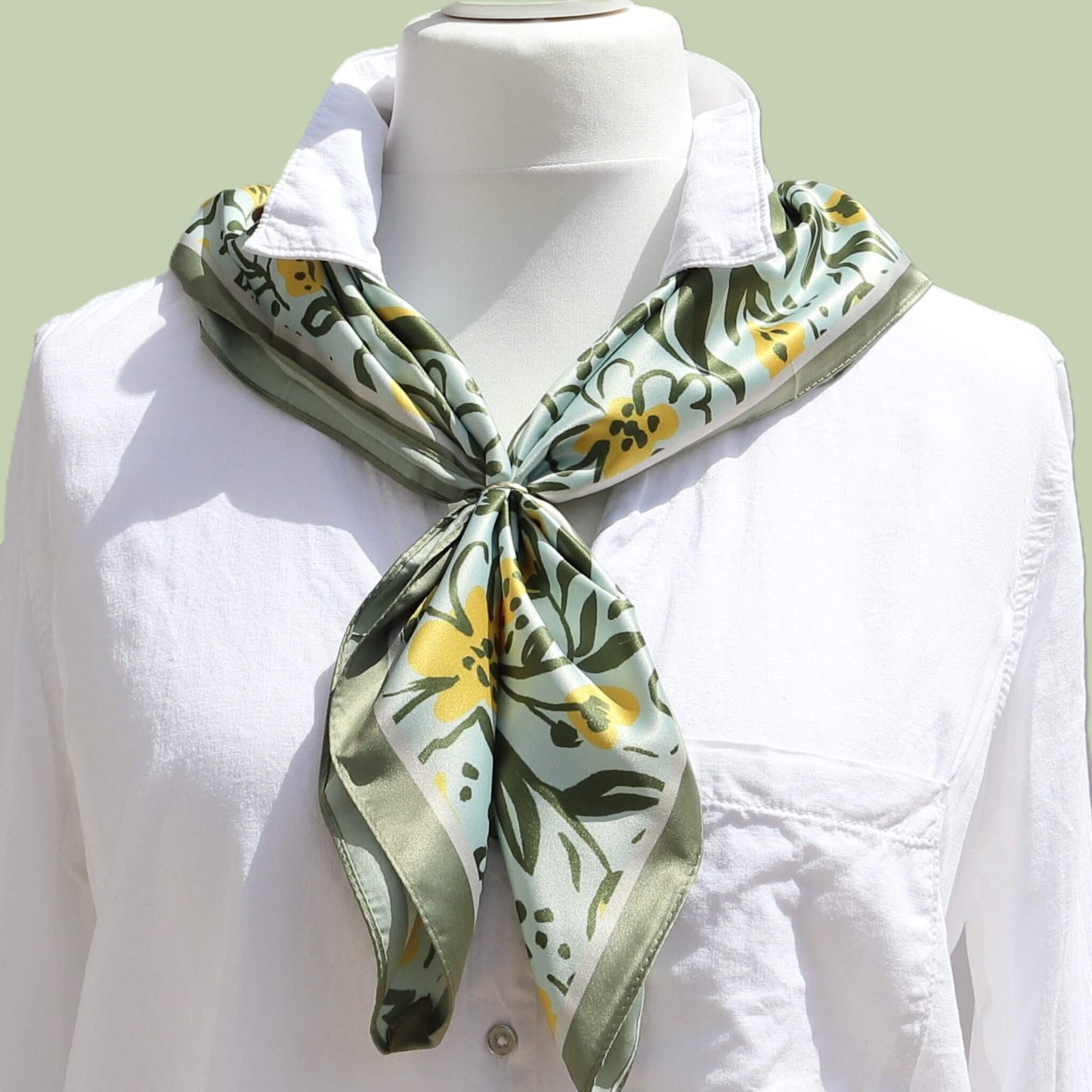 Aurora's Array: Small square silk scarf with a floral pattern in shades of green and yellow on a light blue background, tied loosely around the neck of a mannequin wearing a white shirt.