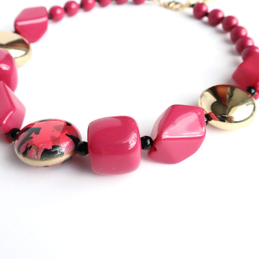Close-up of a short necklace featuring faceted fuchsia beads, shiny gold spheres, and a round bead with a red and black floral design.