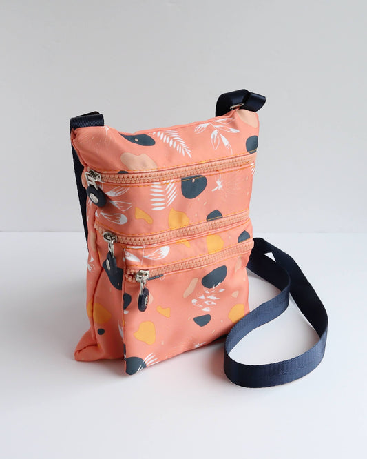 Coral crossbody bag with a navy strap and accents, showcasing a contemporary abstract floral design in navy, yellow, and white. Features three zippered pockets.