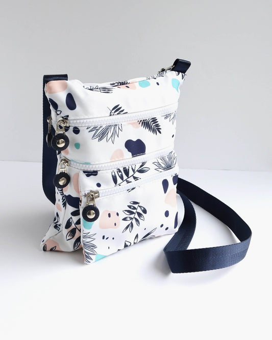 White crossbody bag with a navy strap and trim, featuring a modern abstract floral print in navy, pink, and light blue. Three zippered compartments.