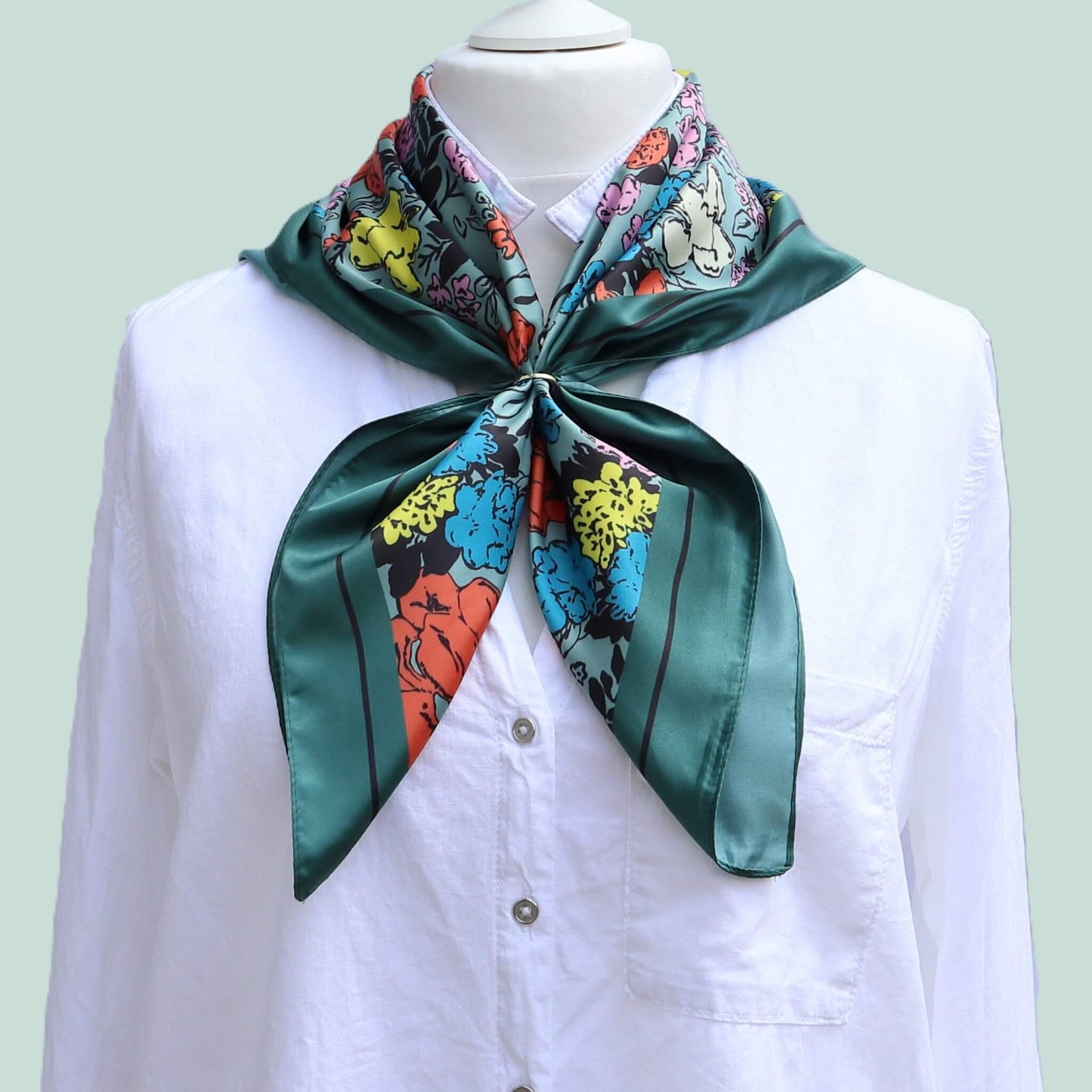 Aurora's Array: The Vibrant Floral Satin Square Scarf - A dark teal satin scarf with a bold floral print in shades of pink, yellow, blue, and orange, tied neatly around the neck of a mannequin wearing a white collared shirt.