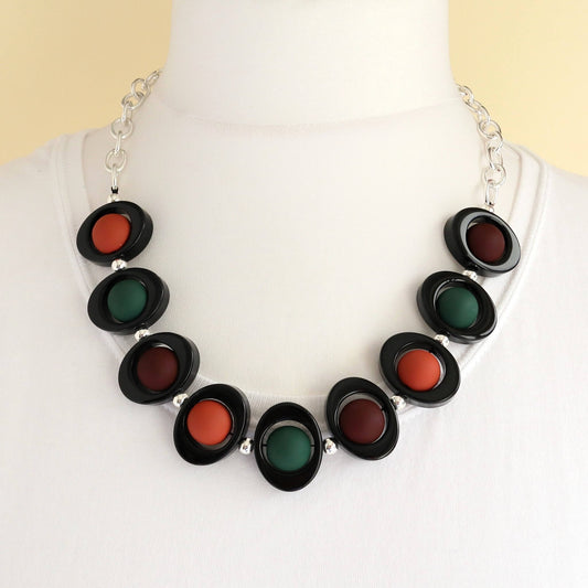 Modern chunky necklace with black oval links enclosing matte burnt orange, dark green, and deep red circular beads. Silver-toned chain.
