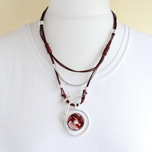 Contemporary layered statement necklace with deep red cords and silver-tone details. Features a circular pendant with a red and white marbled centre.