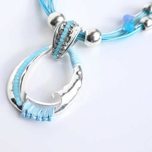 Silver abstract oval pendant necklace with teal cord strands, silver beads, and light blue crystal accents. Features wrapped teal thread.