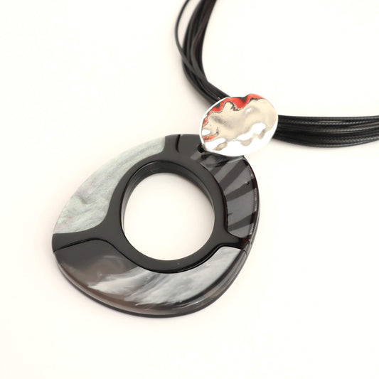 Close-up of a statement necklace with a large, glossy black and grey swirl pendant. Features a smaller, textured silver oval detail and multiple black cords.
