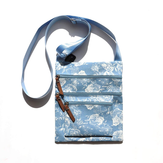 Light blue crossbody bag with a white floral print. Features three zippered sections with brown faux leather pulls and a light blue adjustable strap.