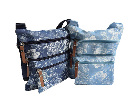 Two floral crossbody bags, one in dark denim with white flowers, the other in light blue with white flowers. Each has three zippered compartments and an adjustable strap.