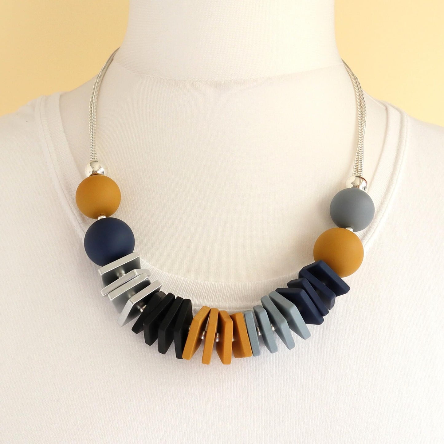 Chunky statement necklace with mustard yellow, navy blue, and grey round beads. Features geometric silver, black, orange, and blue flat beads on a silver chain.