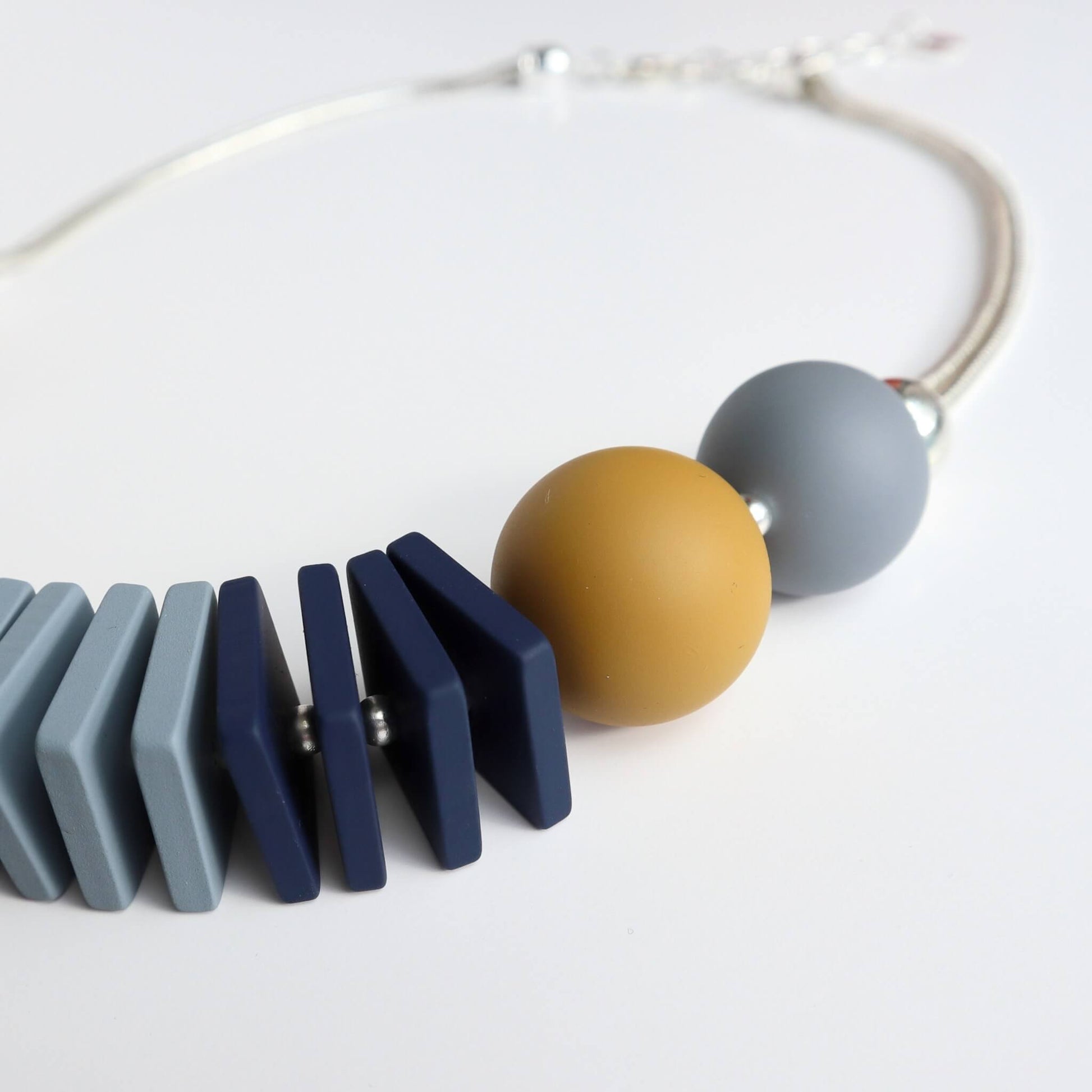 Close-up of a chunky necklace featuring mustard yellow and grey round beads, and geometric flat beads in shades of blue and grey on a silver chain.