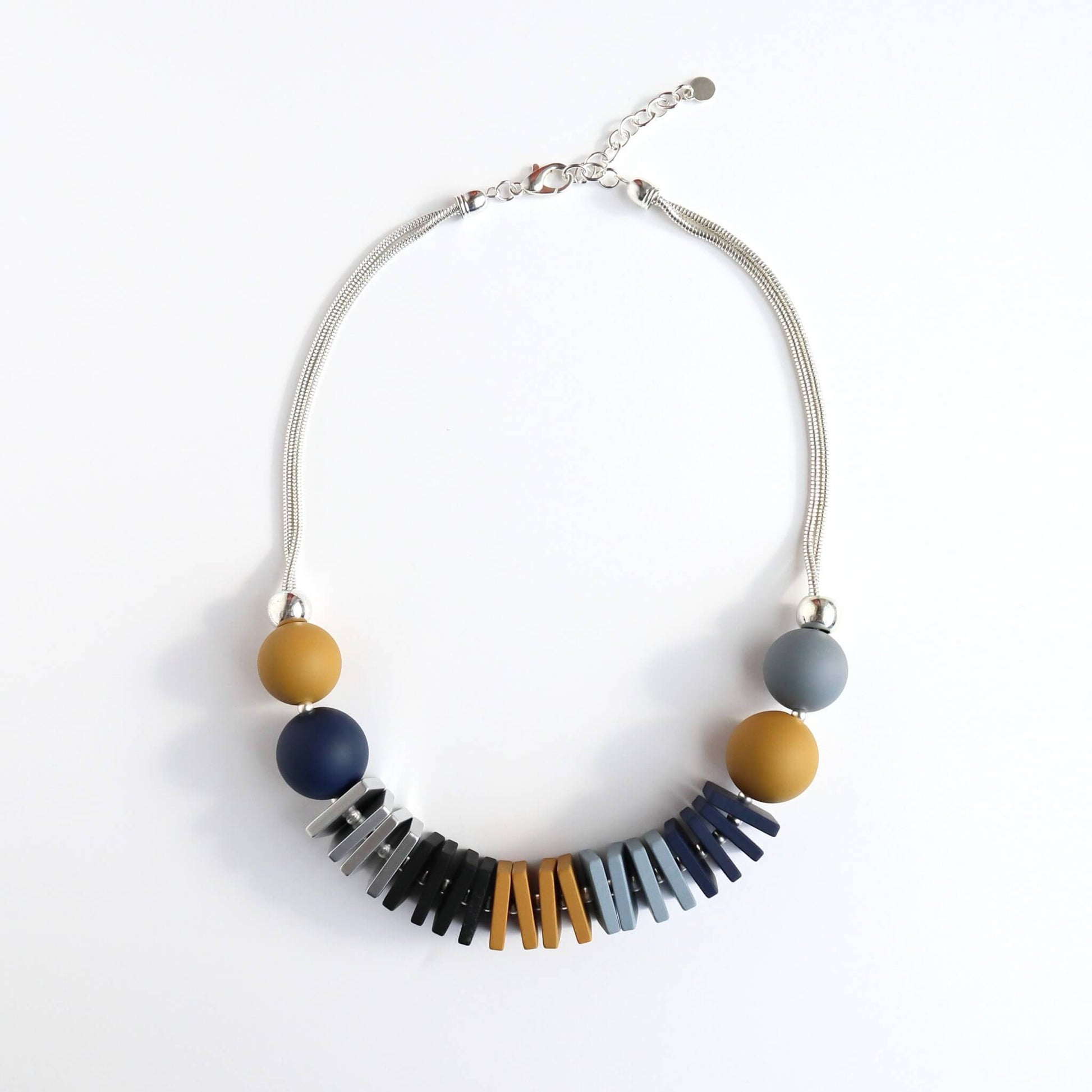 Statement necklace with mustard yellow, navy, and grey round beads. Features silver, black, orange, and blue geometric flat beads on a silver-toned chain with an adjustable clasp.