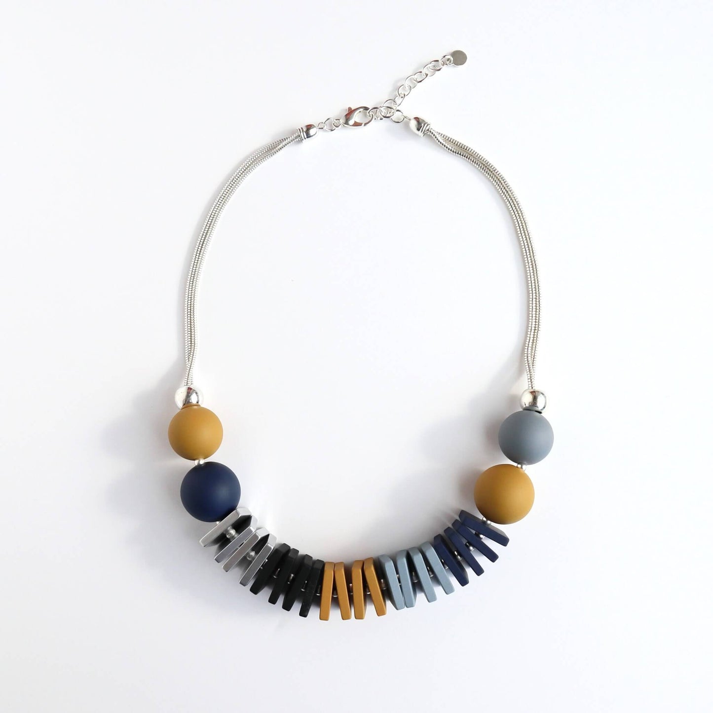 Statement necklace with mustard yellow, navy, and grey round beads. Features silver, black, orange, and blue geometric flat beads on a silver-toned chain with an adjustable clasp.