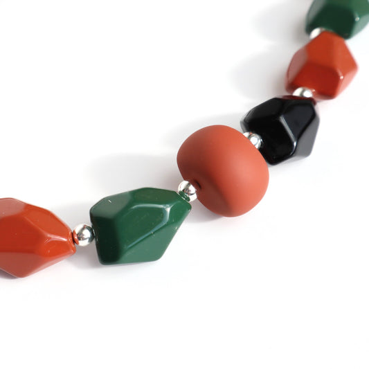 Close-up of a short necklace featuring matte rust-orange, shiny black, and glossy dark green irregular beads, separated by small silver spacers.