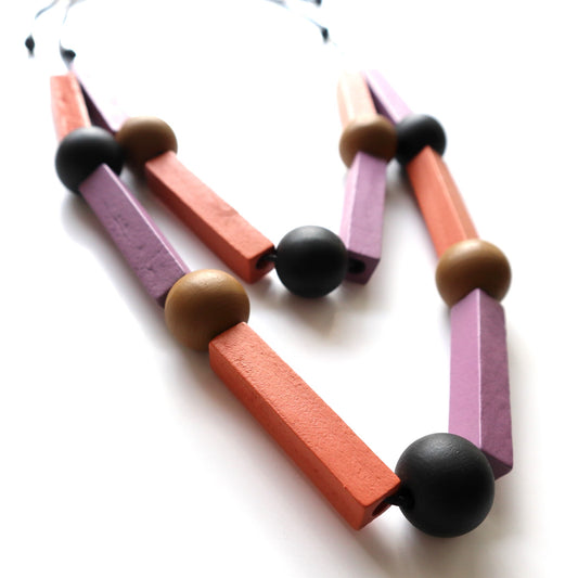 Geometric statement necklace with a black cord, featuring matte lavender and orange rectangular wooden beads, and smooth tan and black round wooden beads.