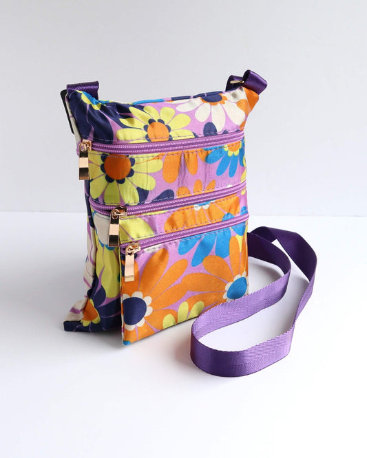 Colorful floral crossbody bag with a purple strap and trim, featuring daisies in orange, yellow, blue, and purple. Three zippered compartments with gold pulls.