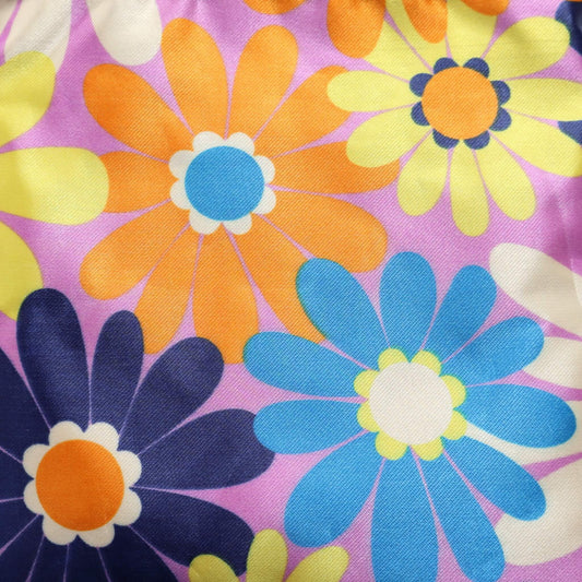 Close-up of a crossbody bag's fabric: a vibrant daisy print in shades of purple, orange, yellow, blue, and white. Detailed floral design.