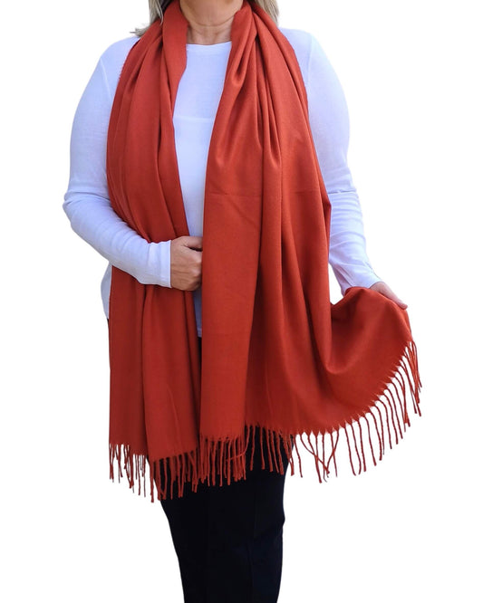 Aurora's Array: Woman models a soft, warm rust-colored wool blend scarf with fringed edges, draped around her neck and shoulders.