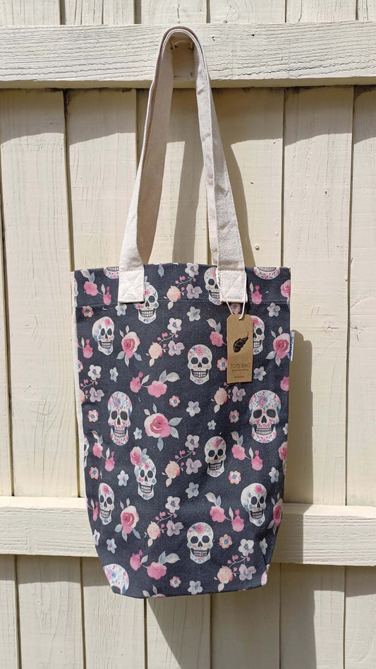 Dark grey canvas tote featuring a print of skulls and pink roses. Includes an interior pocket and sturdy cream-colored carrying straps.