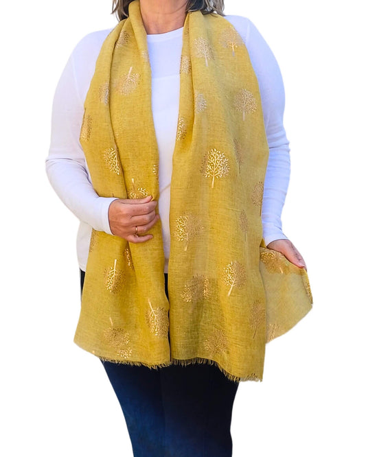 Aurora's Array: Woman models a soft, mustard yellow scarf with a subtle rose gold tree print and frayed edges.