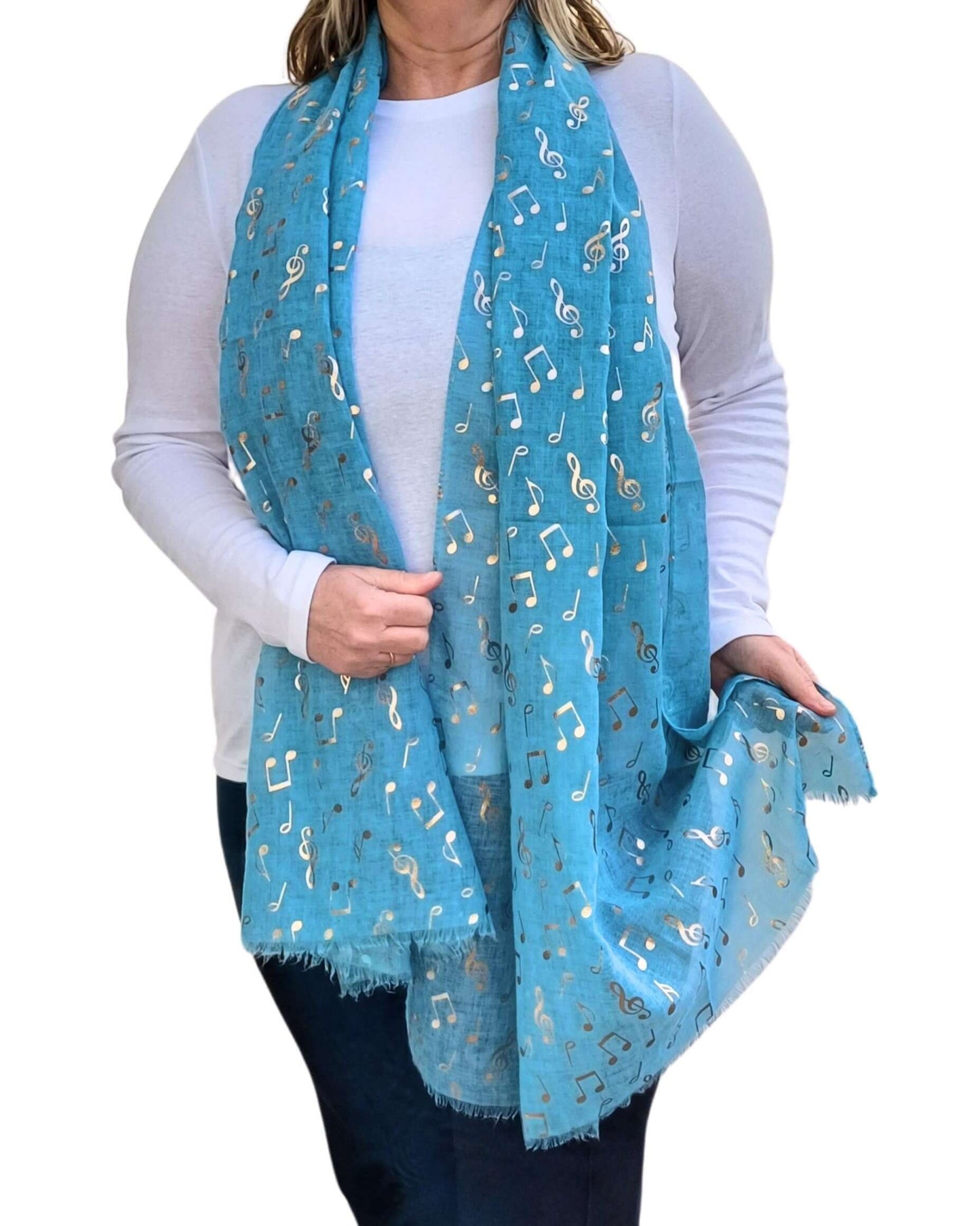 Aurora's Array: A woman wearing a long turquoise scarf featuring metallic rose gold music note prints. The scarf is loosely draped around her neck over a plain white long-sleeve top and paired with dark trousers.