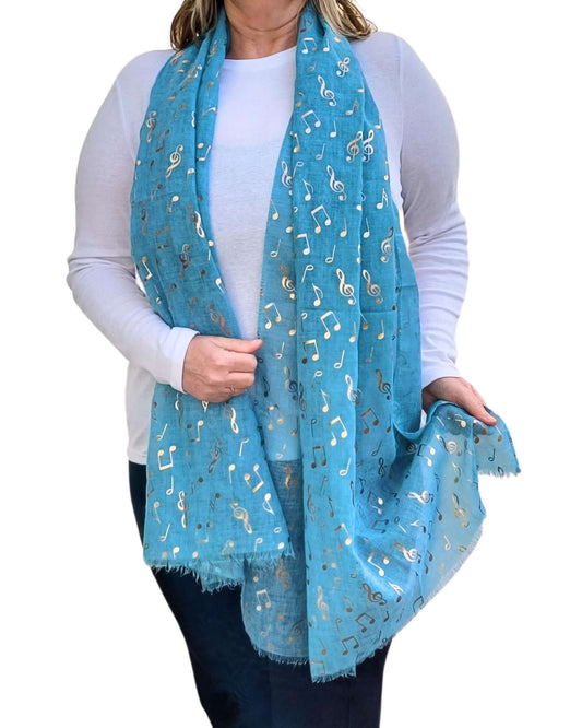 Aurora's Array: A woman wearing a long turquoise scarf featuring metallic rose gold music note prints. The scarf is loosely draped around her neck over a plain white long-sleeve top and paired with dark trousers.