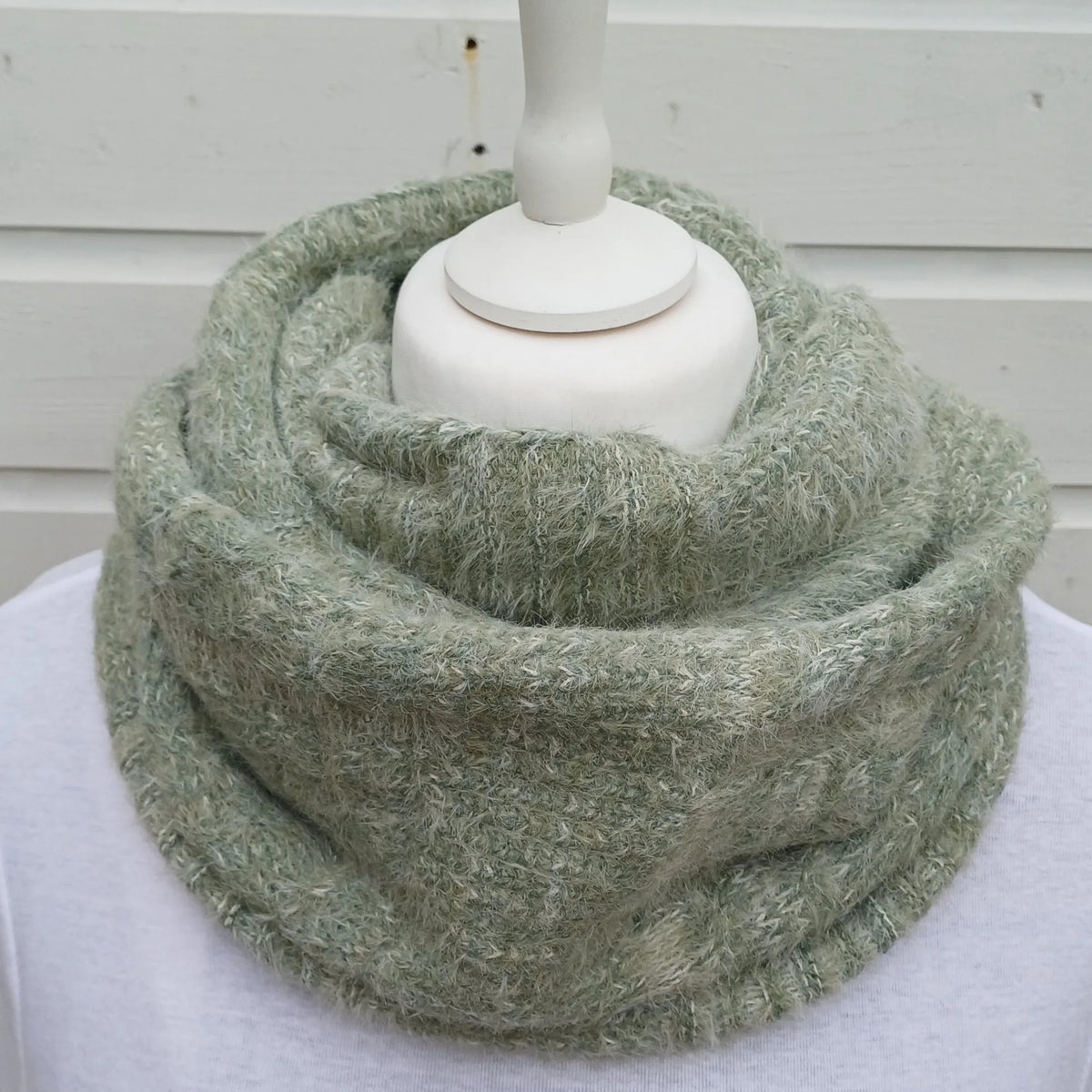 A fuzzy, textured knit infinity scarf in a muted sage green color, displayed on a white mannequin torso against a white wooden background.