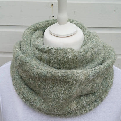 A fuzzy, textured knit infinity scarf in a muted sage green color, displayed on a white mannequin torso against a white wooden background.