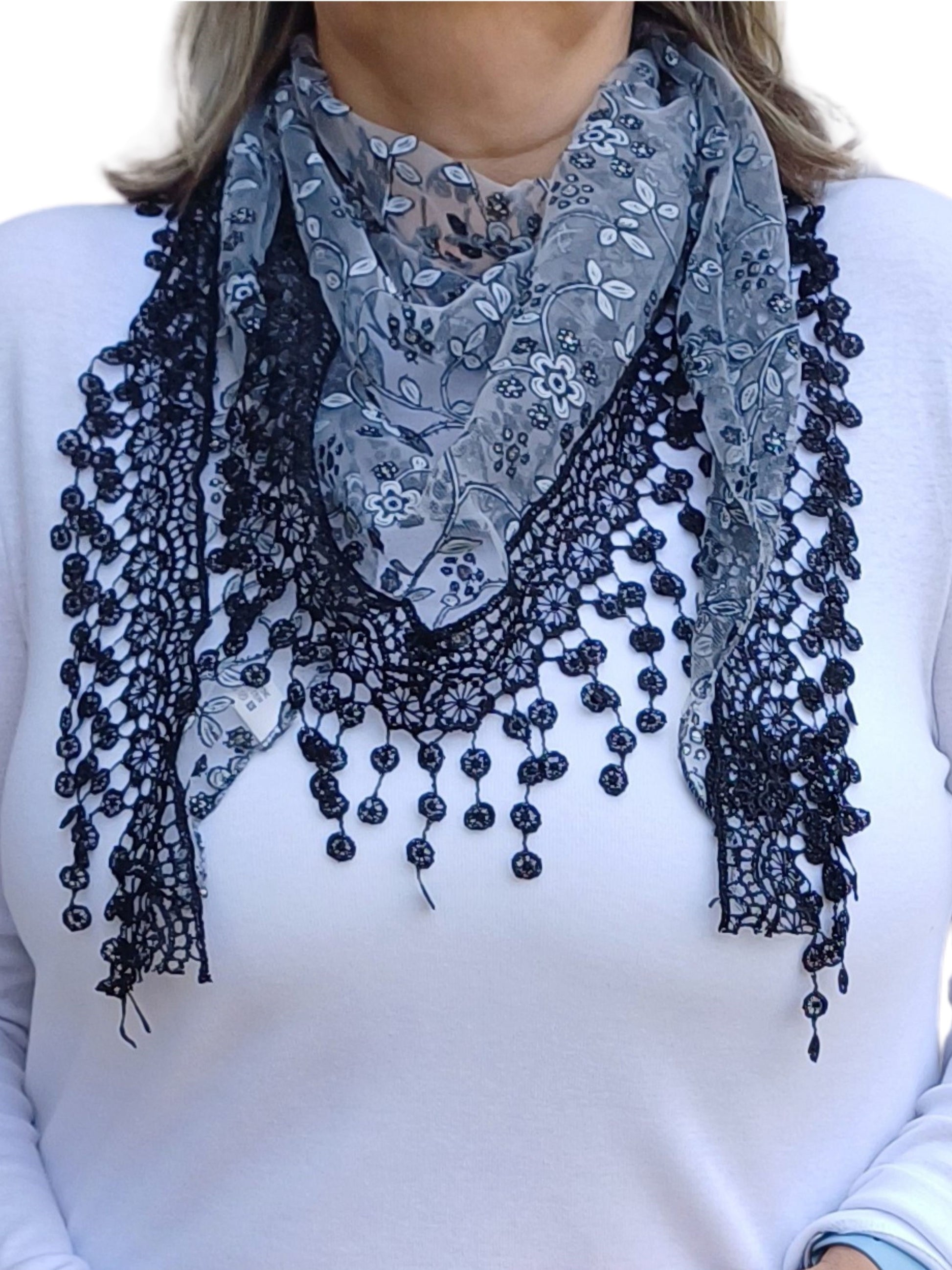 Aurora's Array Glitter Triangle Scarf with Lace & Embroidery: A woman models a grey, sheer triangle scarf with a delicate white floral print, intricate black lace trim, and dangling embroidered accents.