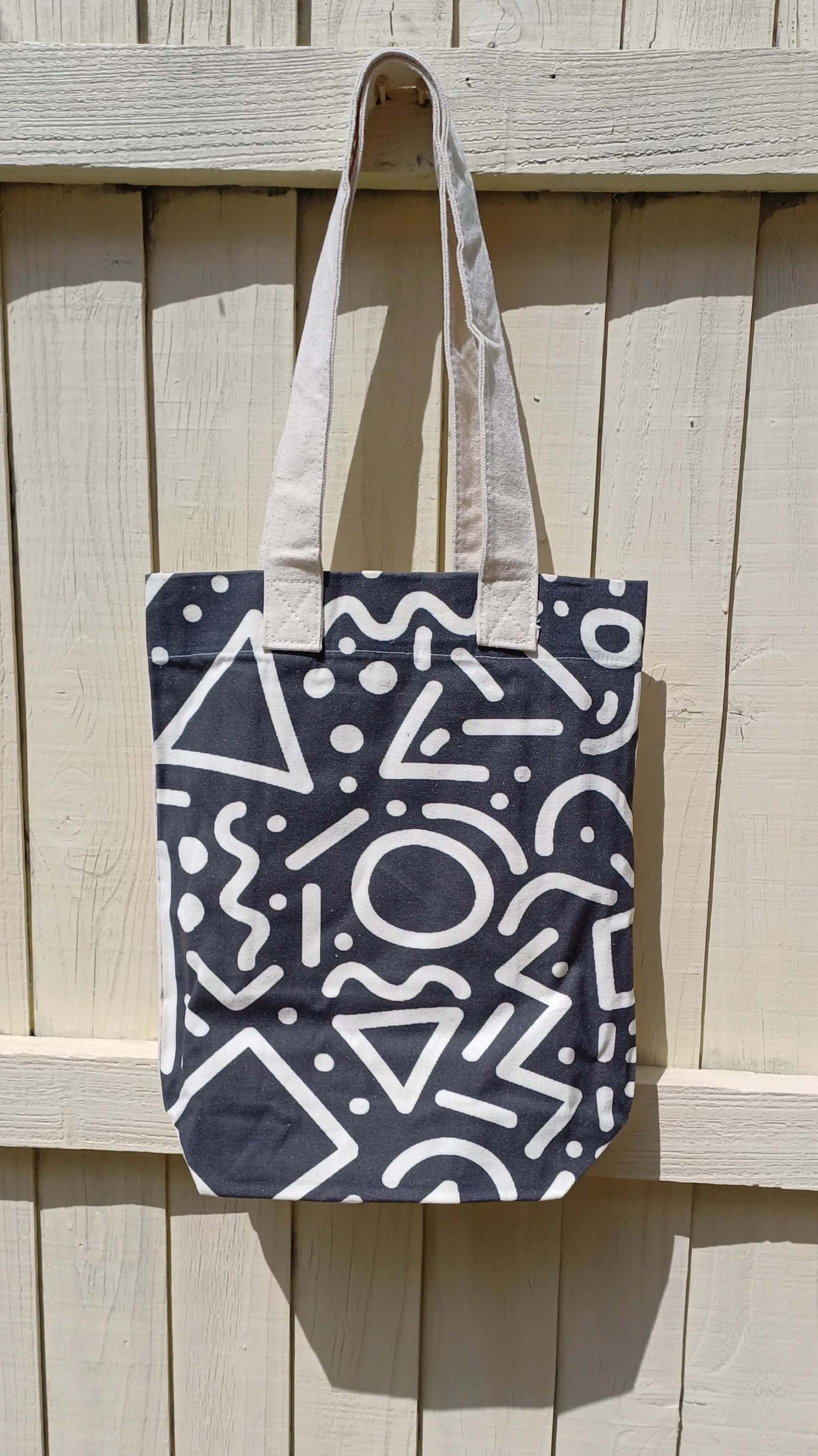 Black canvas tote bag featuring a bold white graffiti doodle print. Includes a convenient inner pocket and sturdy cream-colored handles.