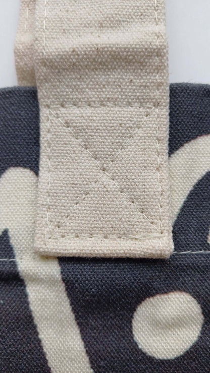Close-up of a cream-colored canvas handle securely stitched with white thread to a black graffiti-print tote bag. Durable fabric detail.