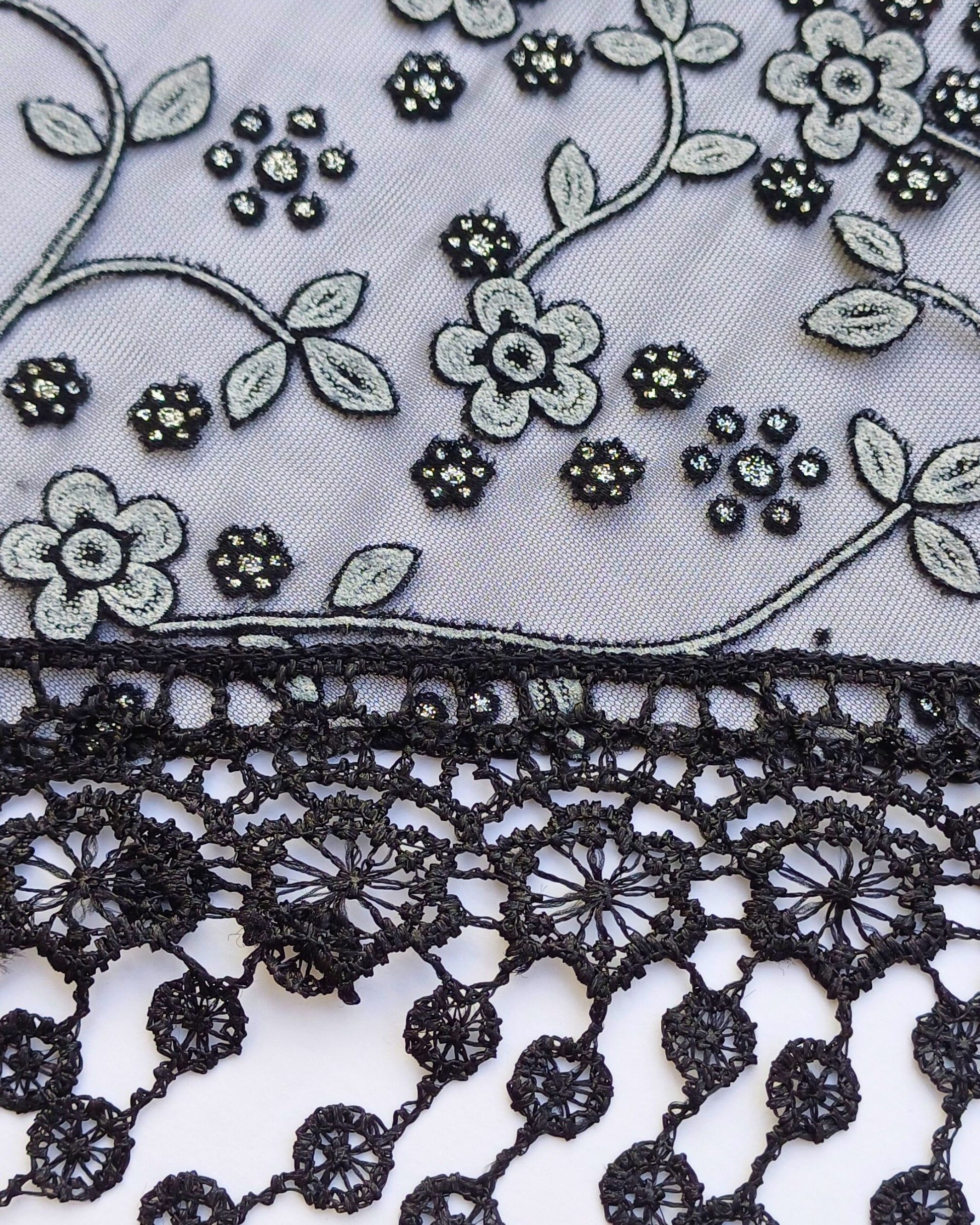 Aurora's Array Glitter Triangle Scarf with Lace & Embroidery (Close-up): A detailed close-up of a white, sheer fabric with delicate grey floral embroidery accented with subtle glitter. The edge features intricate black lace trim with dangling embroidered 