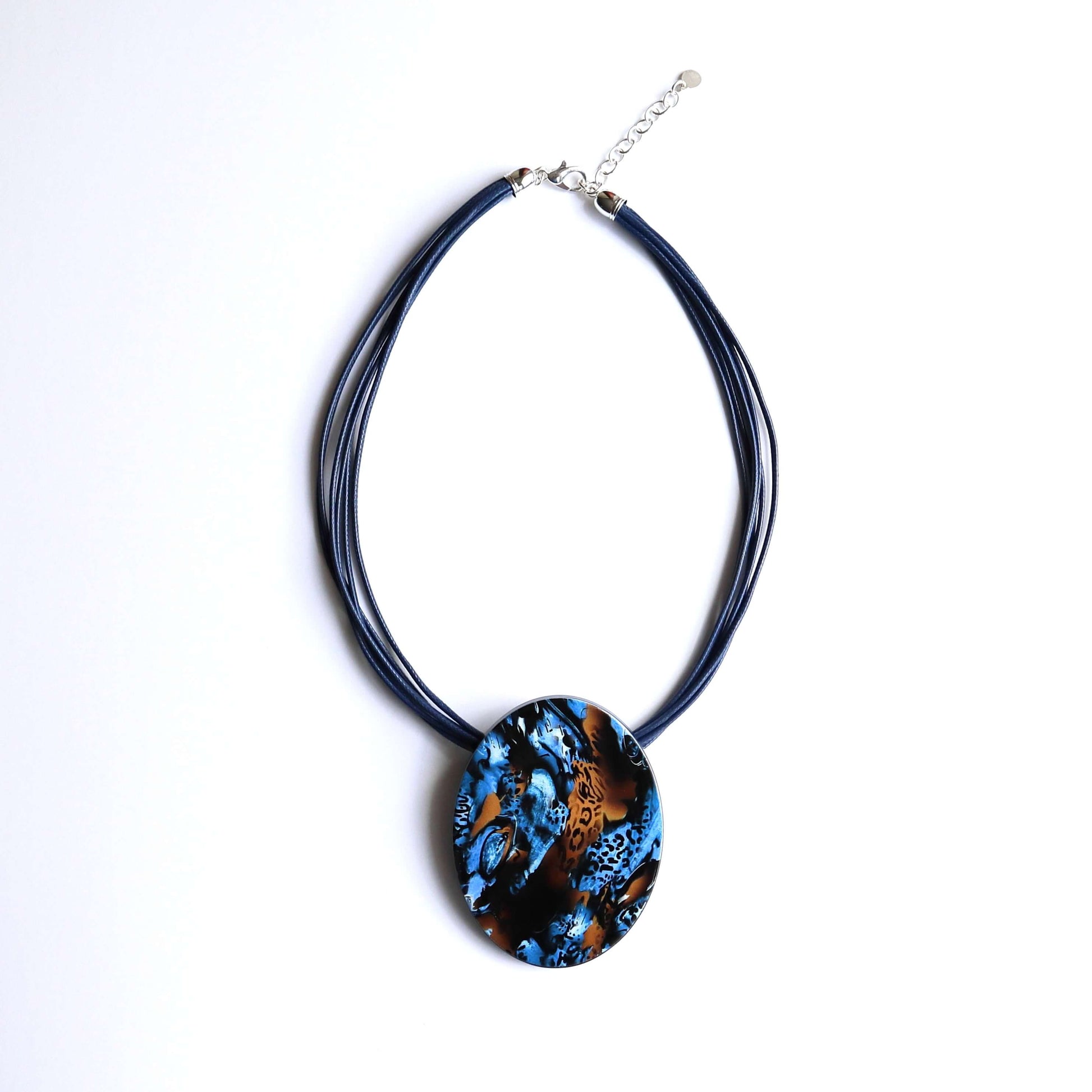 Oval pendant necklace with a vibrant abstract pattern in blue, black, and orange animal print. Suspended from multiple navy blue cords with a silver clasp.