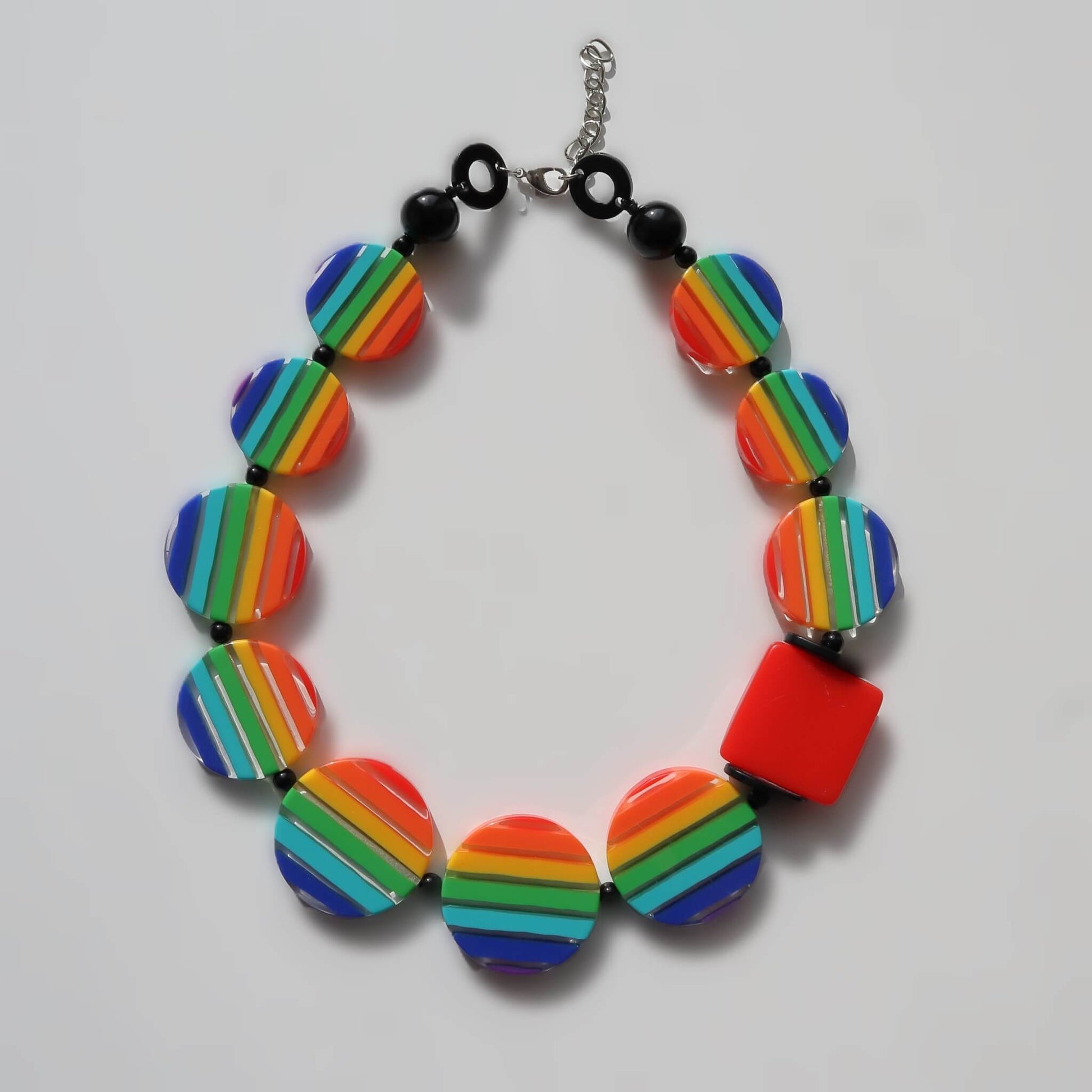 Chunky statement necklace with vibrant rainbow striped round beads and a red square bead. Features black spacer beads and a silver-toned chain.