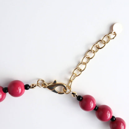 Close-up of a gold-toned necklace clasp and extender chain. Features a lobster claw closure and round charm at the end. Pink and black beads are visible.