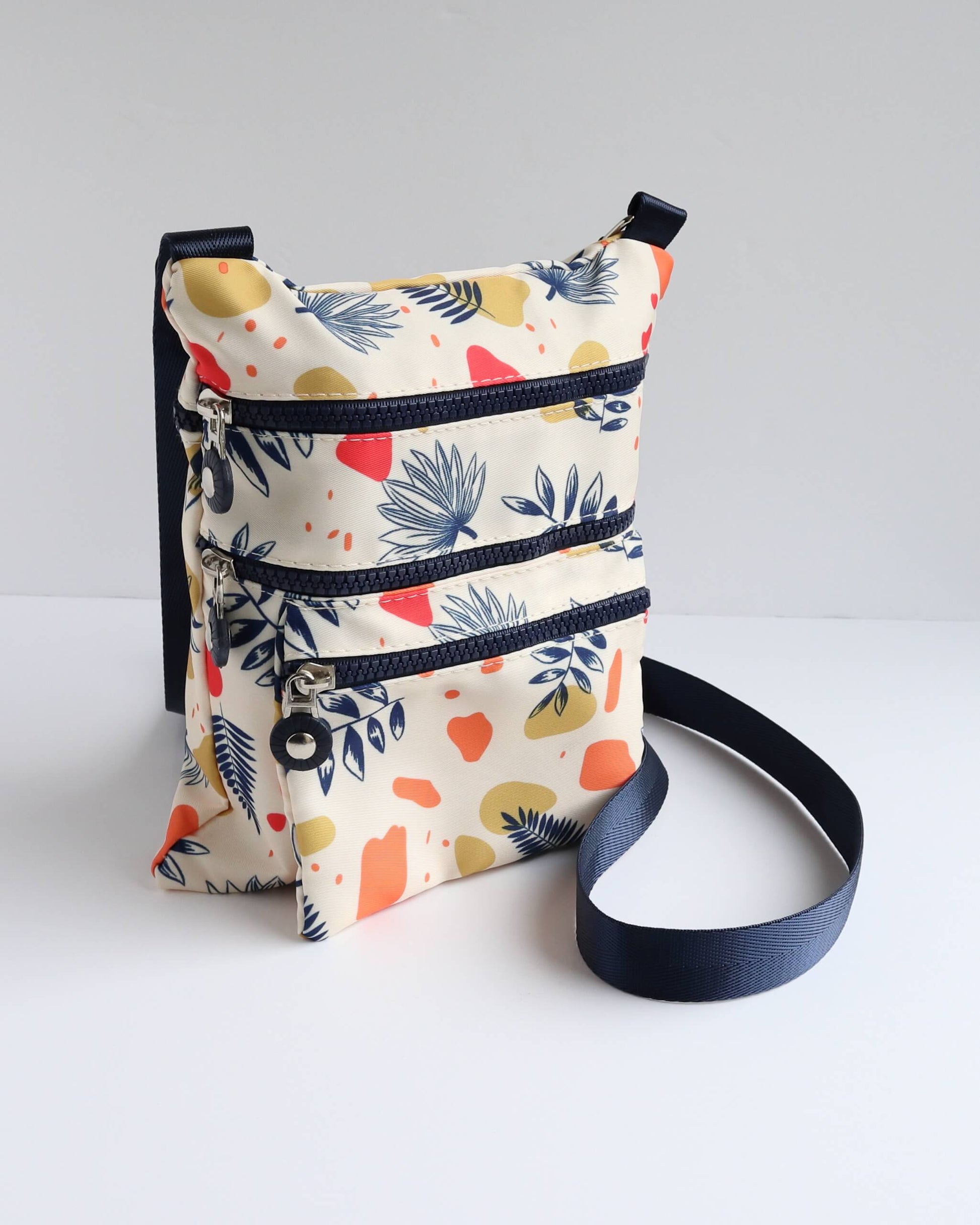 Cream crossbody bag with a navy strap and trim, decorated with a modern abstract leaf print in navy, red, and yellow. Three separate zip pockets.