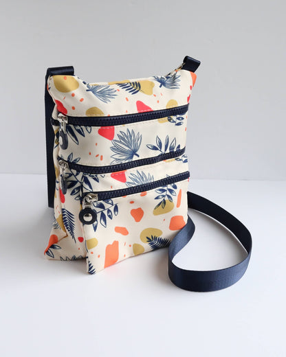 Cream crossbody bag with a navy strap and trim, decorated with a modern abstract leaf print in navy, red, and yellow. Three separate zip pockets.