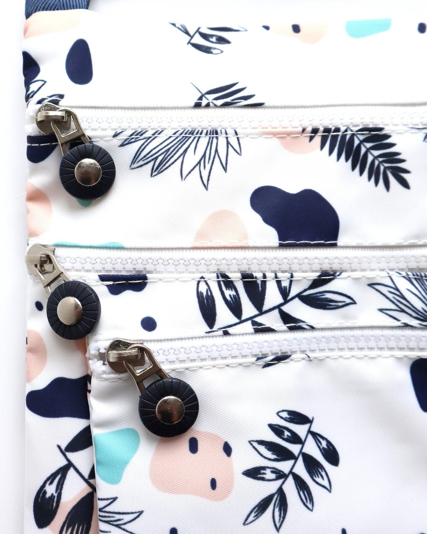 Close-up of a white crossbody bag showing three zippered compartments with silver pulls and black accents. Modern abstract floral print.