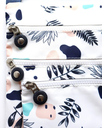 Close-up of a white crossbody bag showing three zippered compartments with silver pulls and black accents. Modern abstract floral print.
