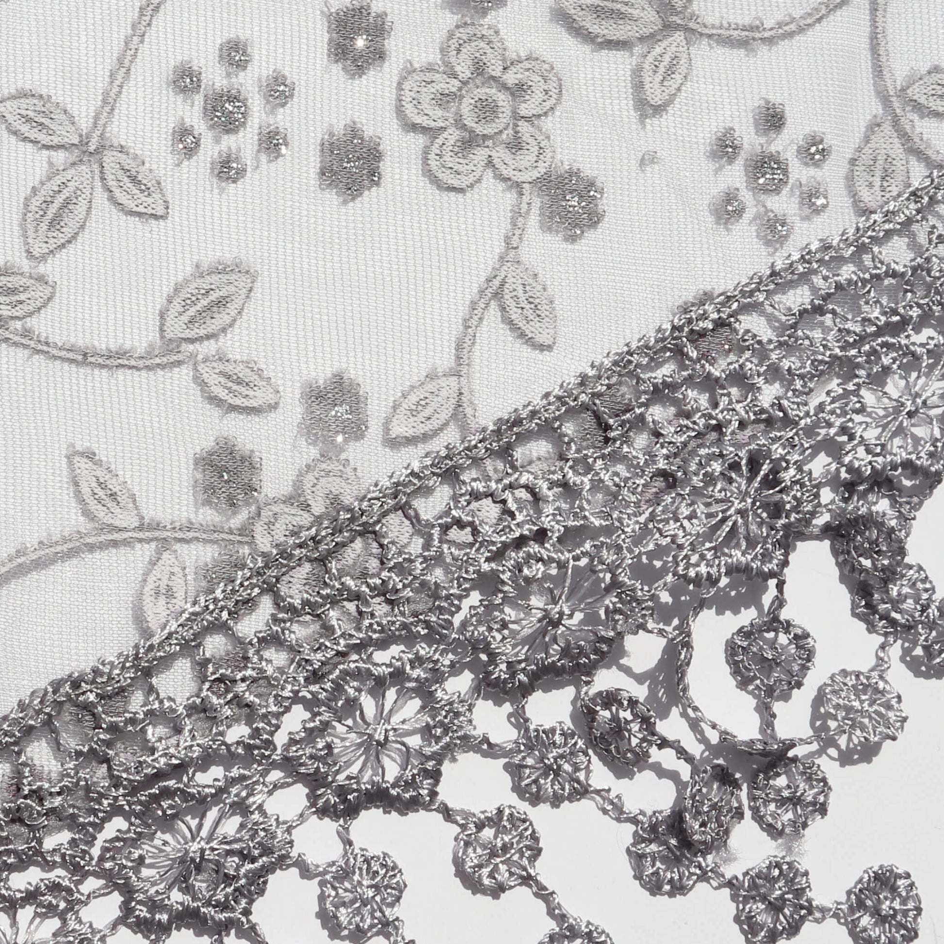 Aurora's Array Glitter Triangle Scarf with Lace & Embroidery (Close-up): A detailed close-up of a white, sheer fabric with delicate grey floral embroidery accented with subtle glitter. The edge features intricate grey lace trim with dangling embroidered d