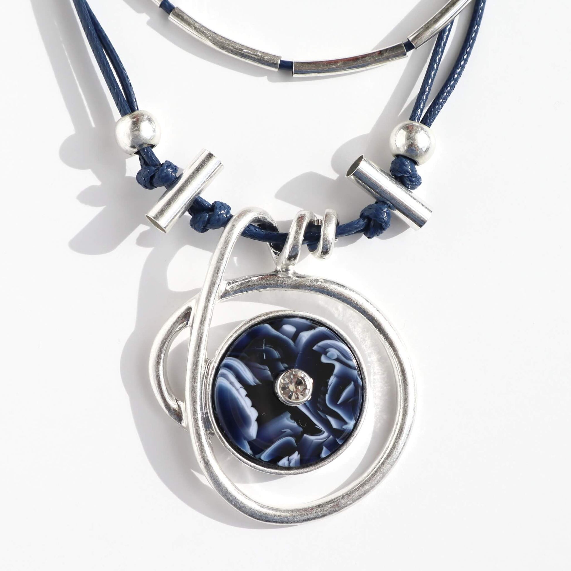 Close-up of a modern layered necklace with navy blue cords and silver-tone accents. Features a circular pendant with a blue and white marbled centre and a small clear stone.